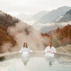 Bassin thermal extérieur fumant dans une vallée espagnole en automne, entouré d’une forêt rousse et de montagnes brumeuses, avec une femme et un homme en peignoirs blancs.